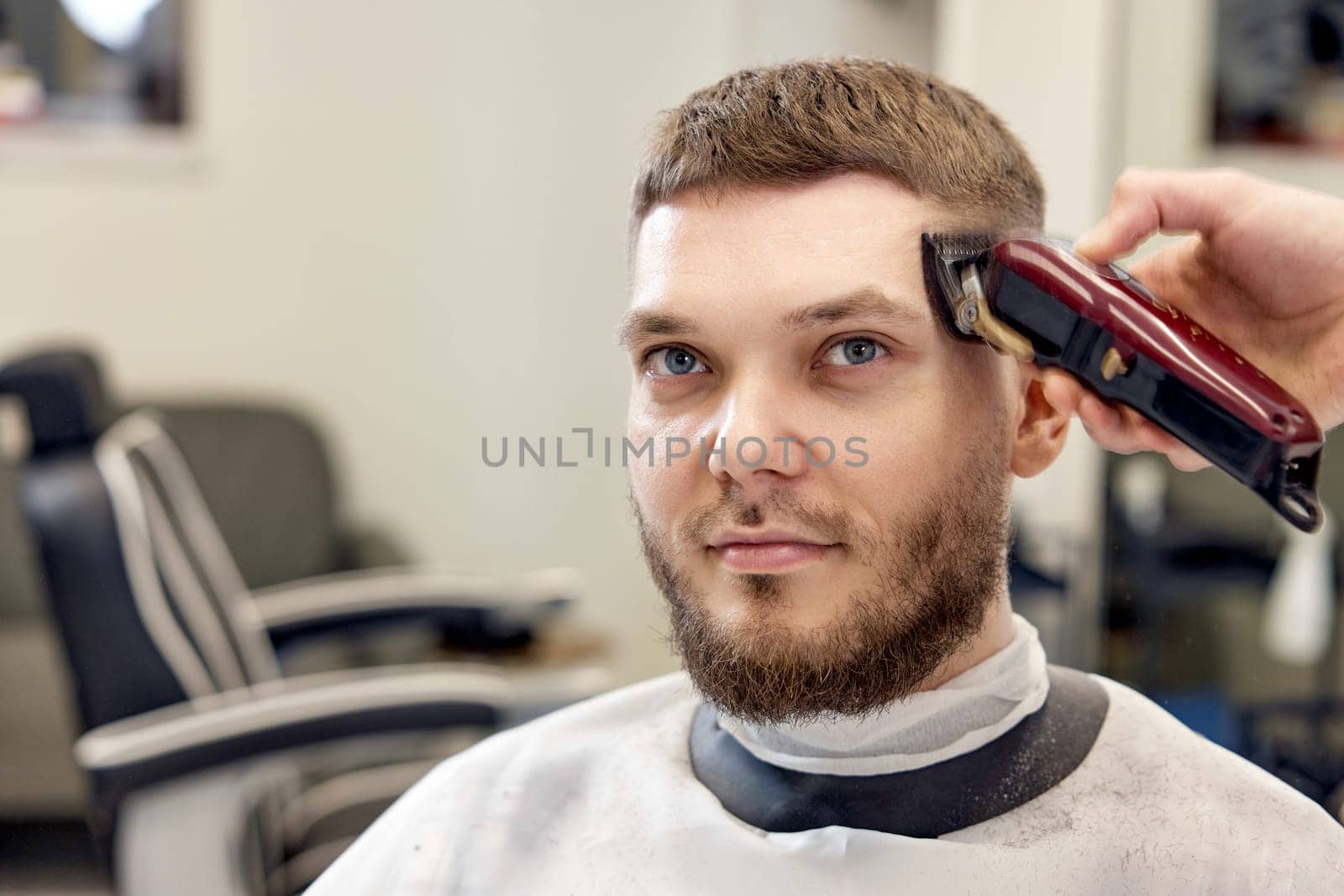 Barber trim hair with clipper on handsome bearded man in barber shop.