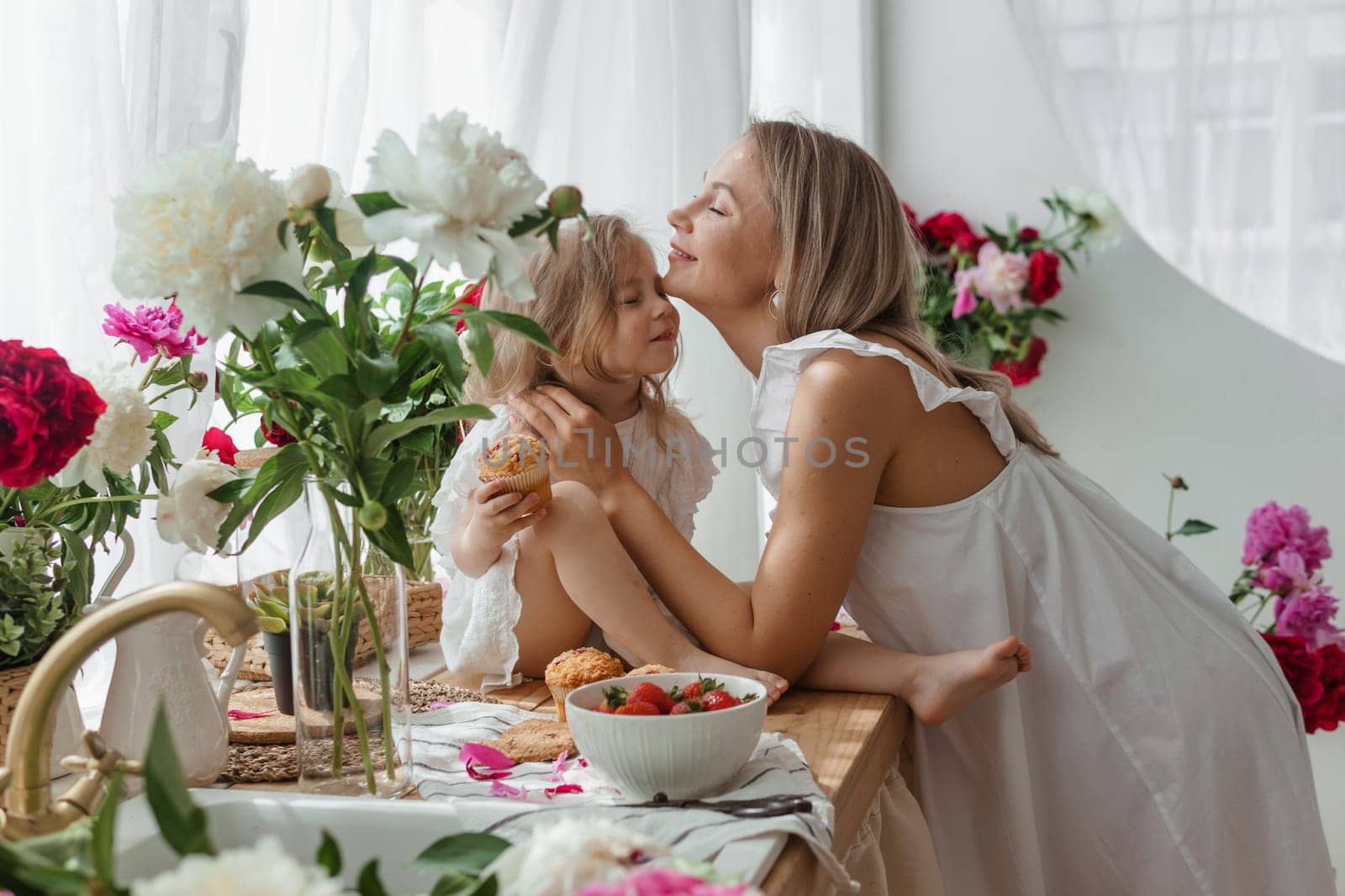 A little blonde girl with her mom on a kitchen countertop decorated with peonies. The concept of the relationship between mother and daughter. Spring atmosphere. by Annu1tochka