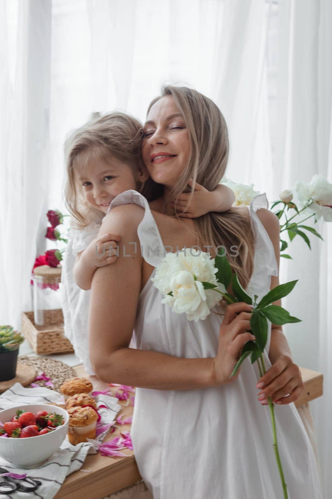 A little blonde girl with her mom on a kitchen countertop decorated with peonies. The concept of the relationship between mother and daughter. Spring atmosphere. by Annu1tochka