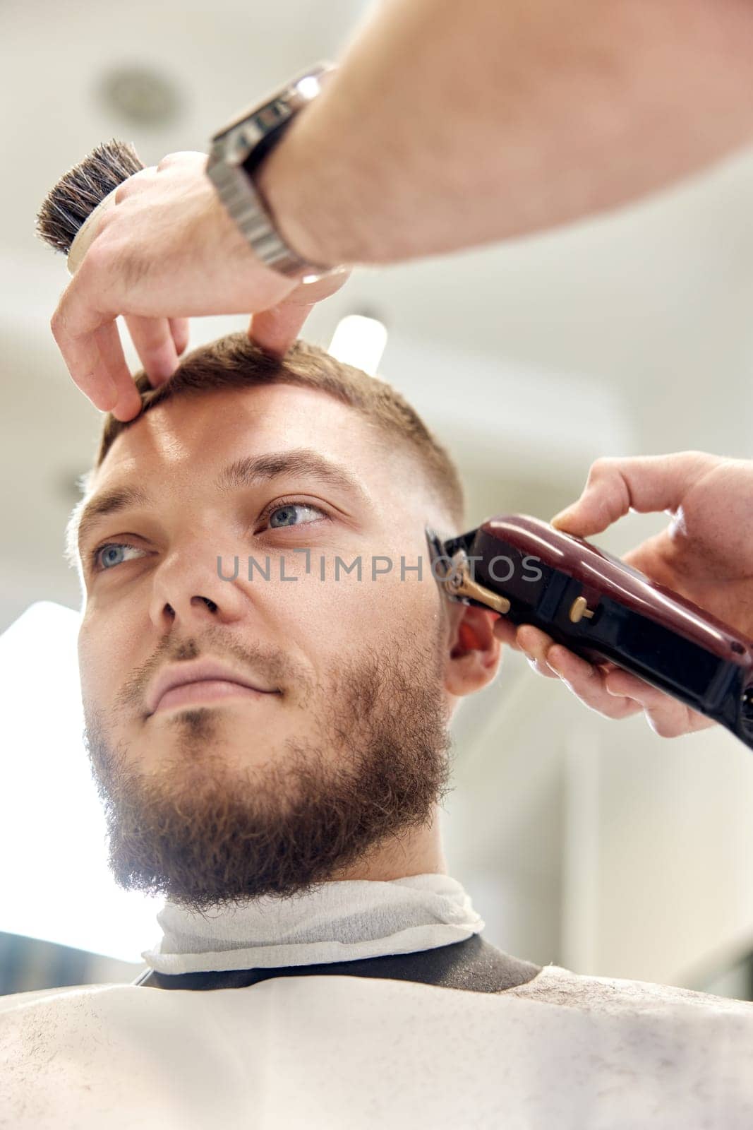 Barber trim hair with clipper on handsome bearded man in barber shop.