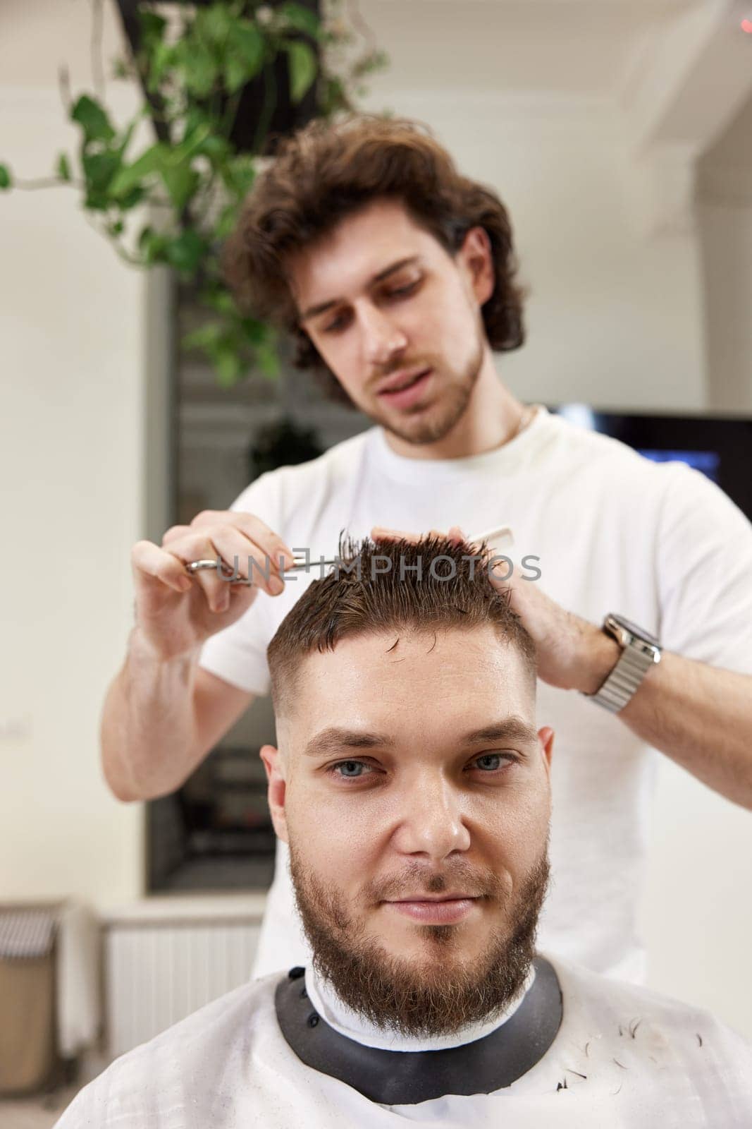 hairdresser does haircut for caucasian bearded man using comb and grooming scissors in barber shop.