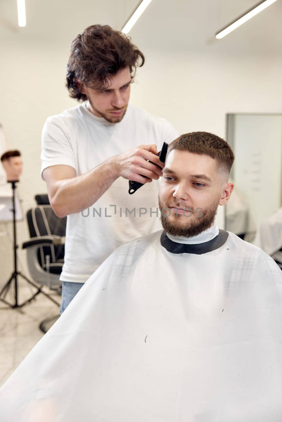 Barber trim hair with clipper on handsome bearded man in barber shop.