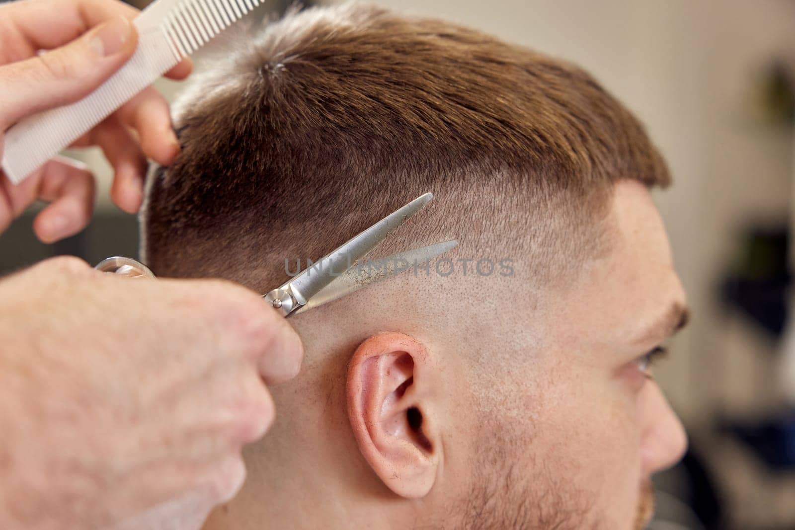 Professional hairdresser does haircut for caucasian bearded man using comb and scissors at barber shop. close-up