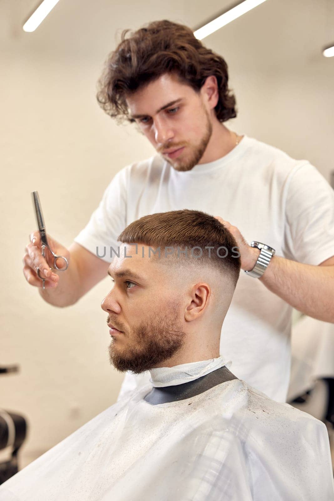 Professional hairdresser does haircut for caucasian bearded man using comb and scissors at barber shop.