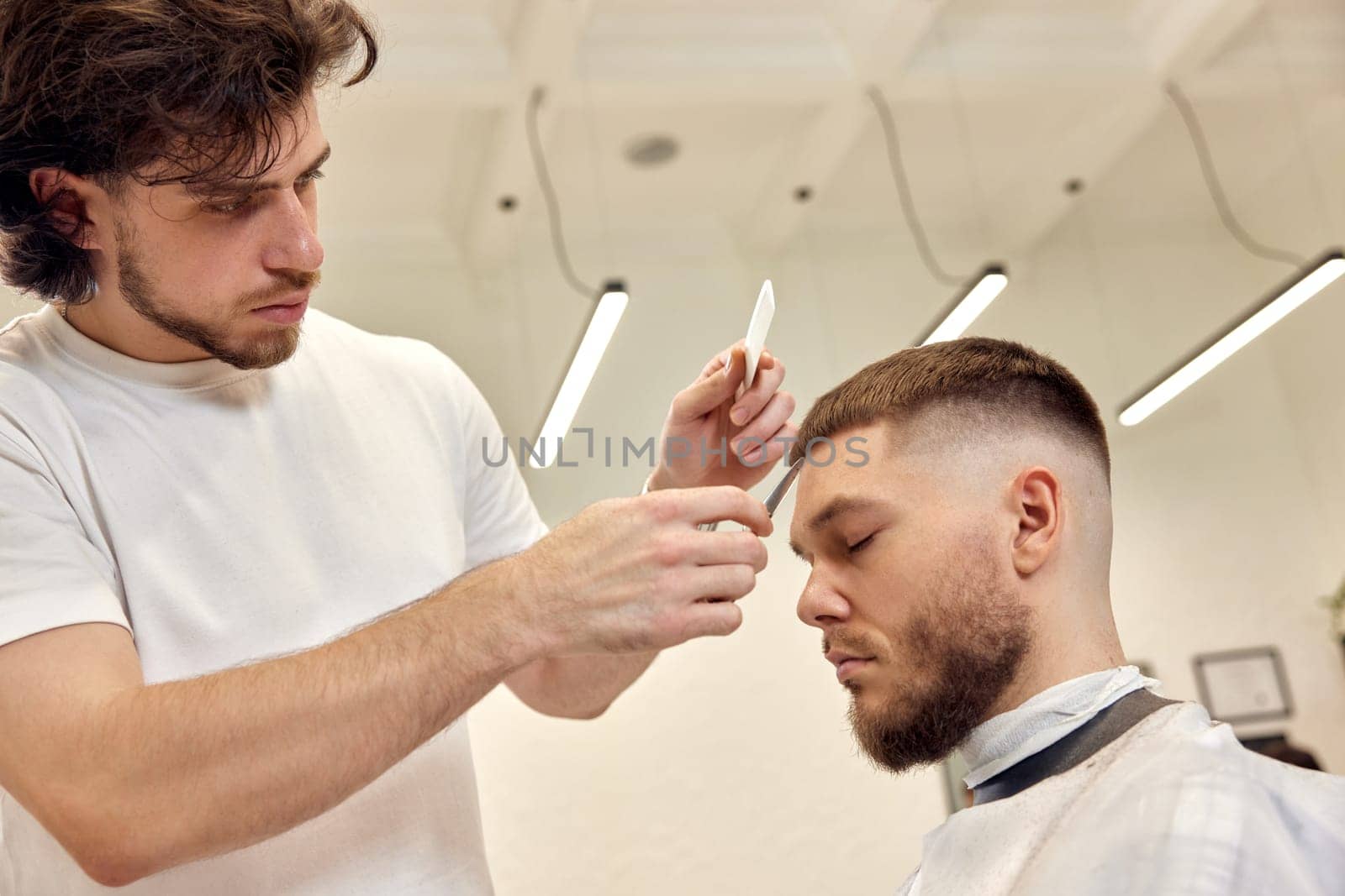 Professional hairdresser does haircut for caucasian bearded man using comb and scissors at barber shop. close-up