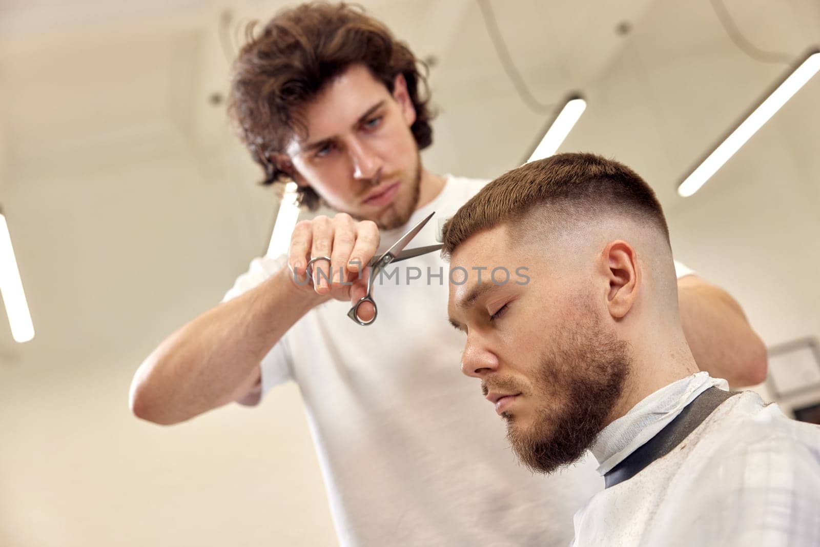 Professional barber does haircut for caucasian bearded man using comb and scissors at barber shop. close-up