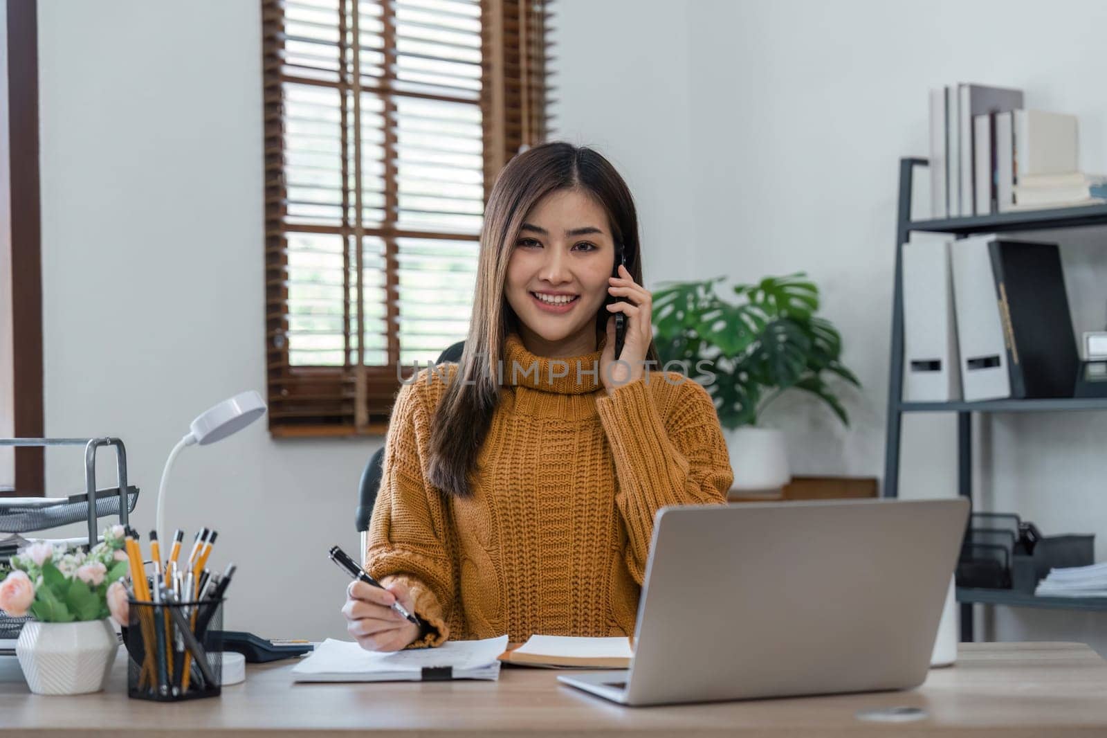 Smiling woman sitting in an home writing down notes talking from her telephone and laptop on the table.