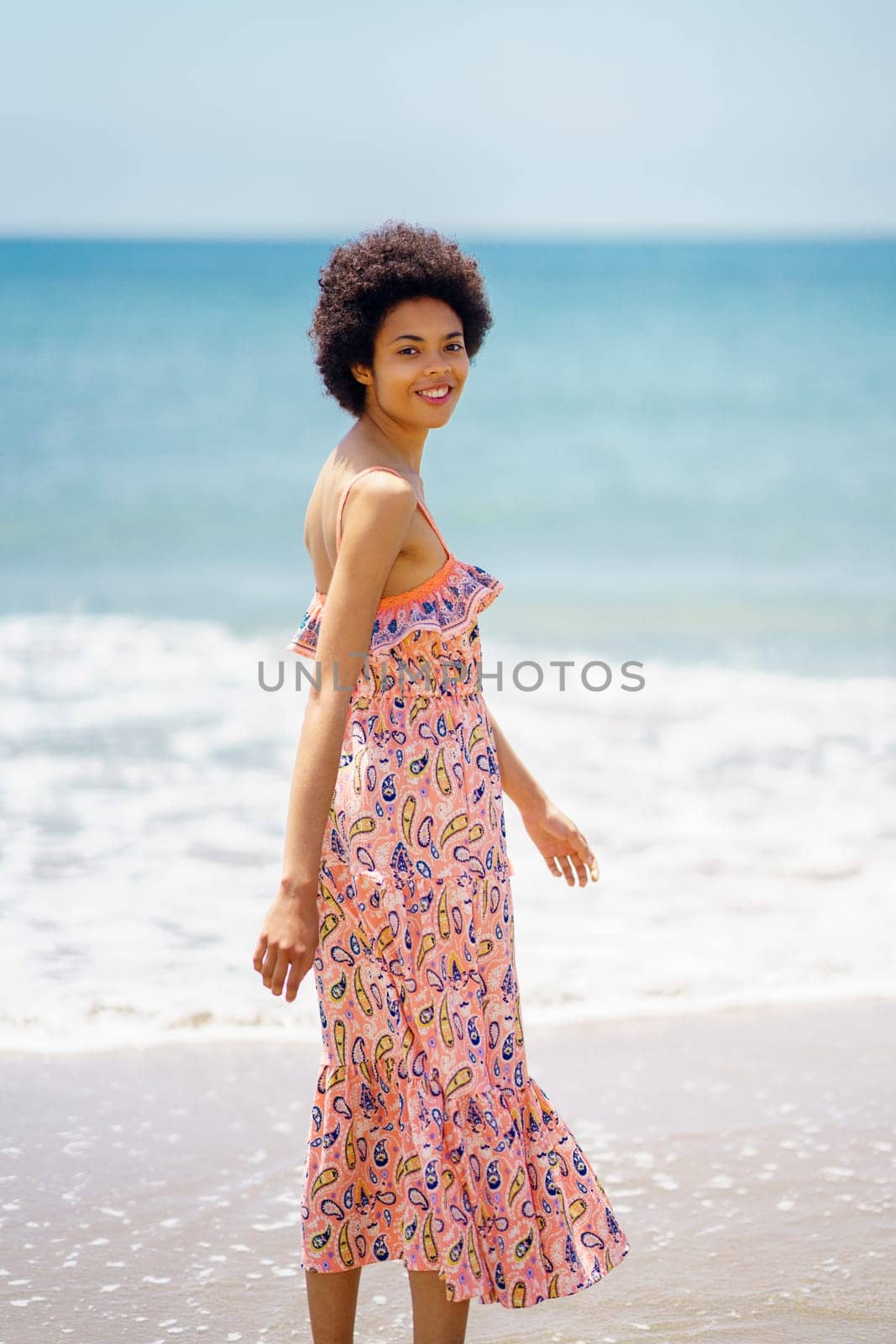 Side view of carefree woman in stylish dress standing on sandy beach washed by rippling sea water and looking at camera