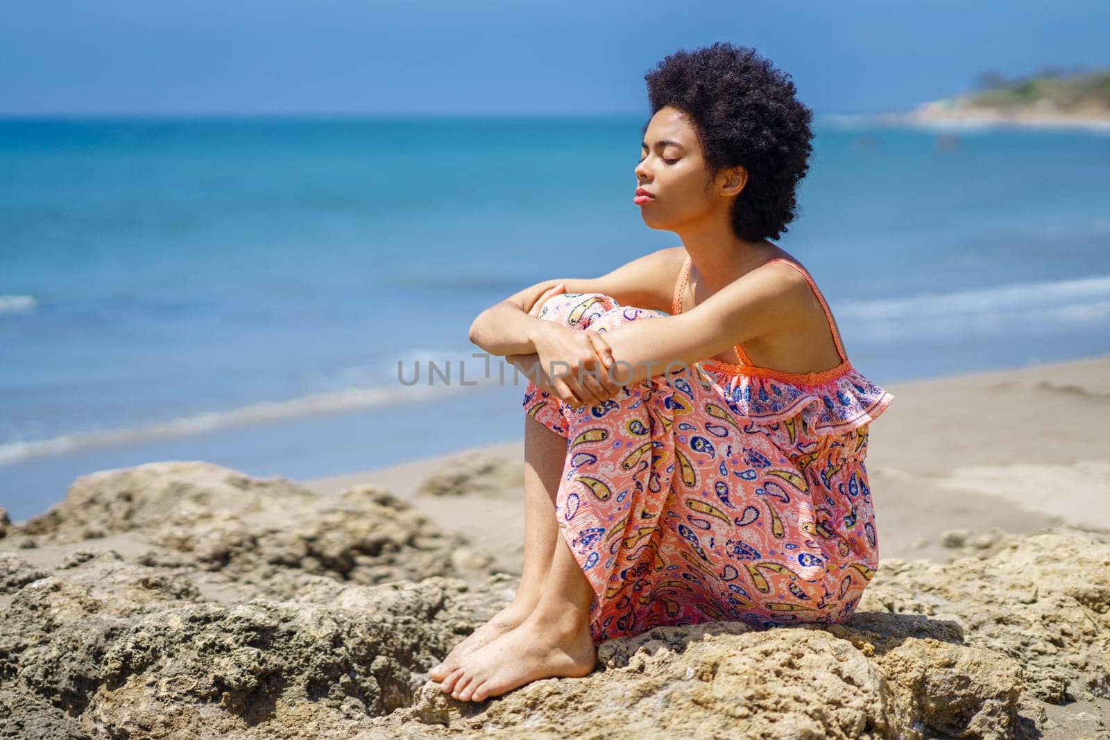 Barefoot black woman in ornamental dress with curly hair, embracing knees and inhaling fresh air with closed eyes while chilling on rough rocks against waving sea on resort