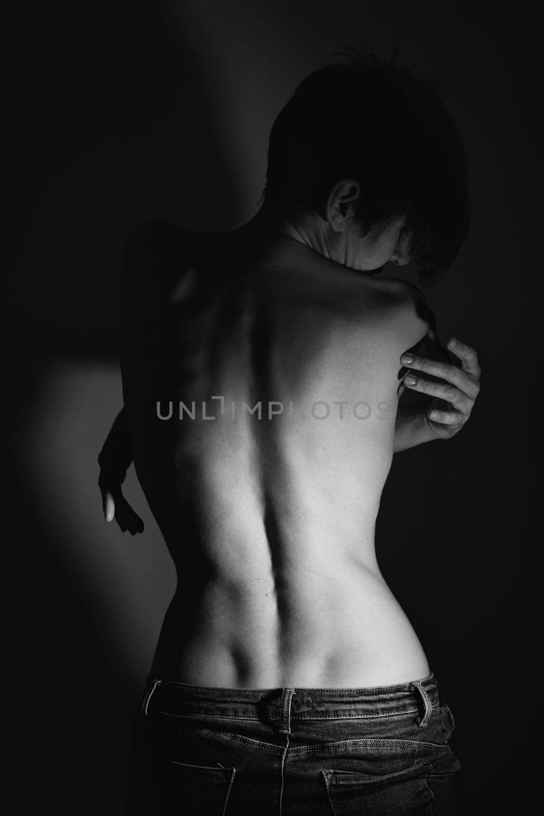 Back view of adult alluring topless woman with short dark hair in jeans standing near wall with crossed arms