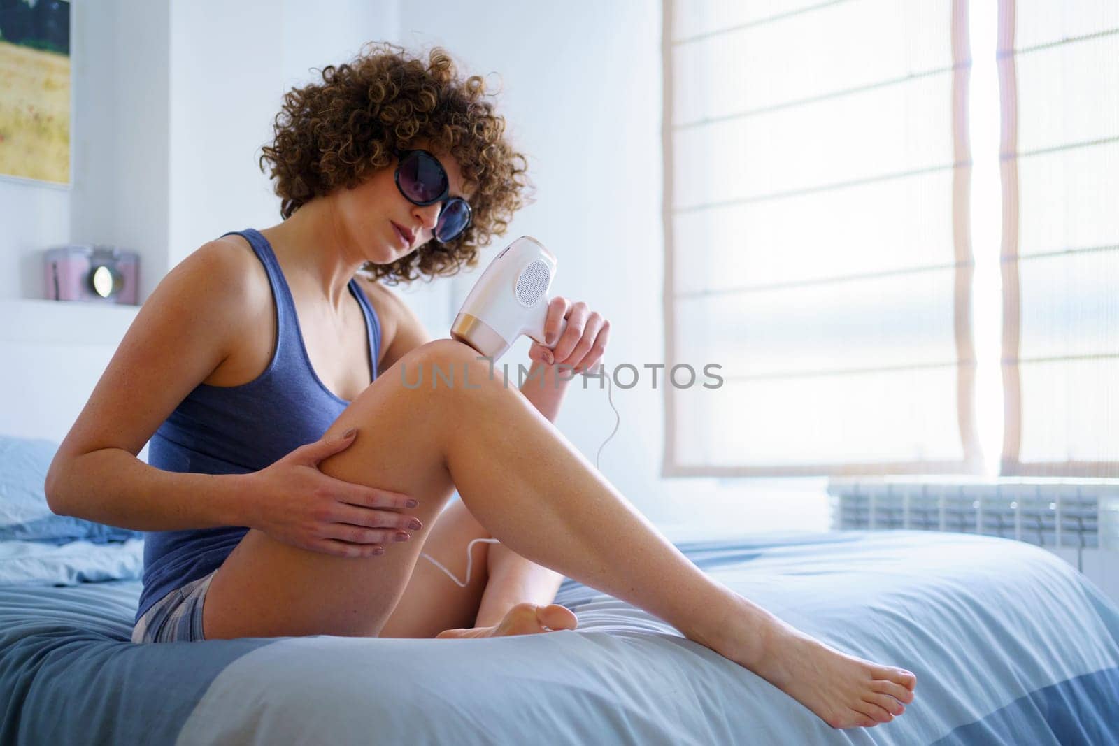 Full body of serious young barefoot female in sleepwear and protective goggles, with Afro hairstyle using IPL for knee hair removal session during skincare routine at home