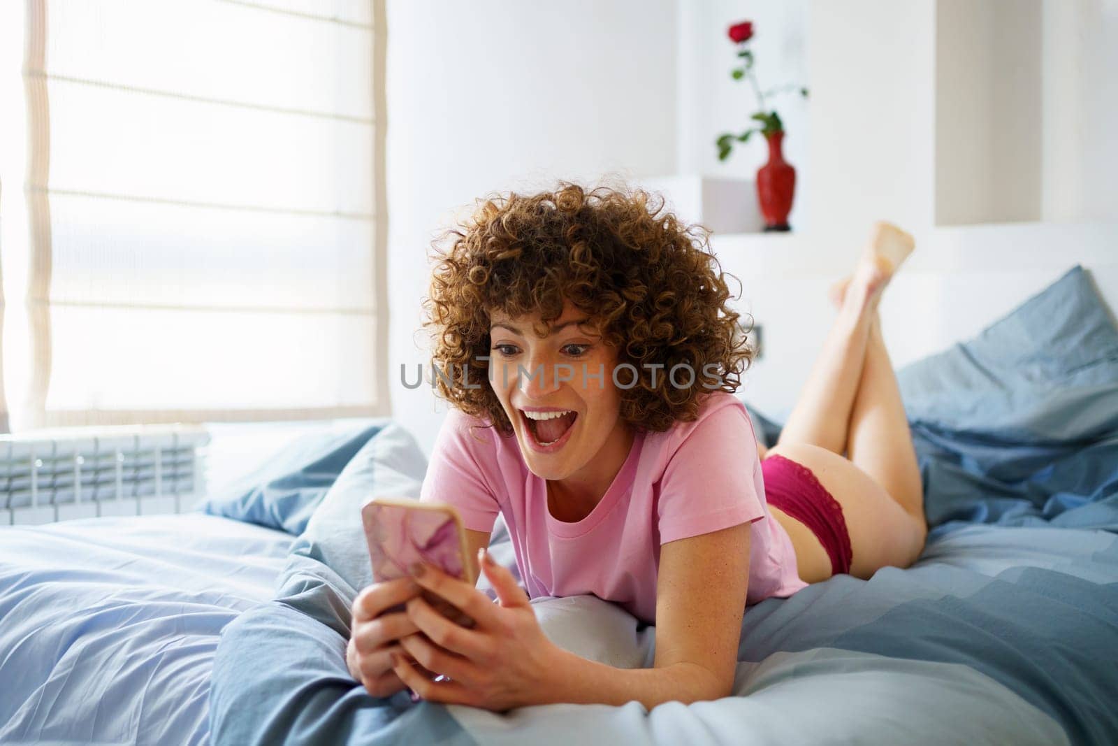 Delighted young woman with curly hair lying on comfortable bed while surfing social media via mobile phone in bedroom at home