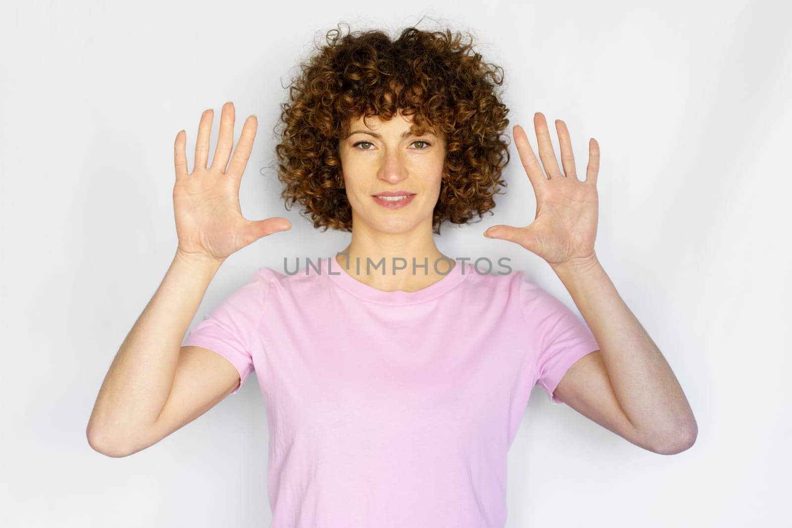 Attractive curly haired girl in casual outfit raising hands and showing number ten while looking at camera on white background