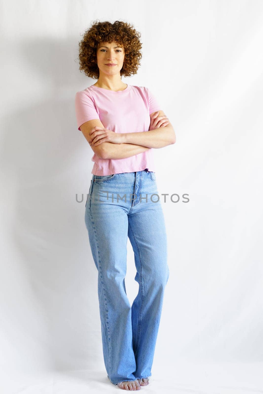Full body of curly haired woman in casual clothes leaning on white wall while standing with hands crossed looking at camera and smiling