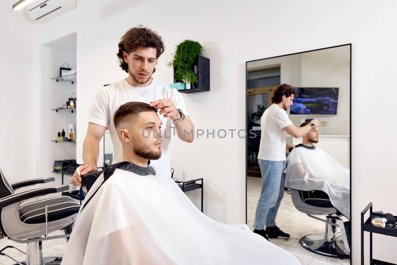 professional male hairstylist combing young customer's hair in barbershop