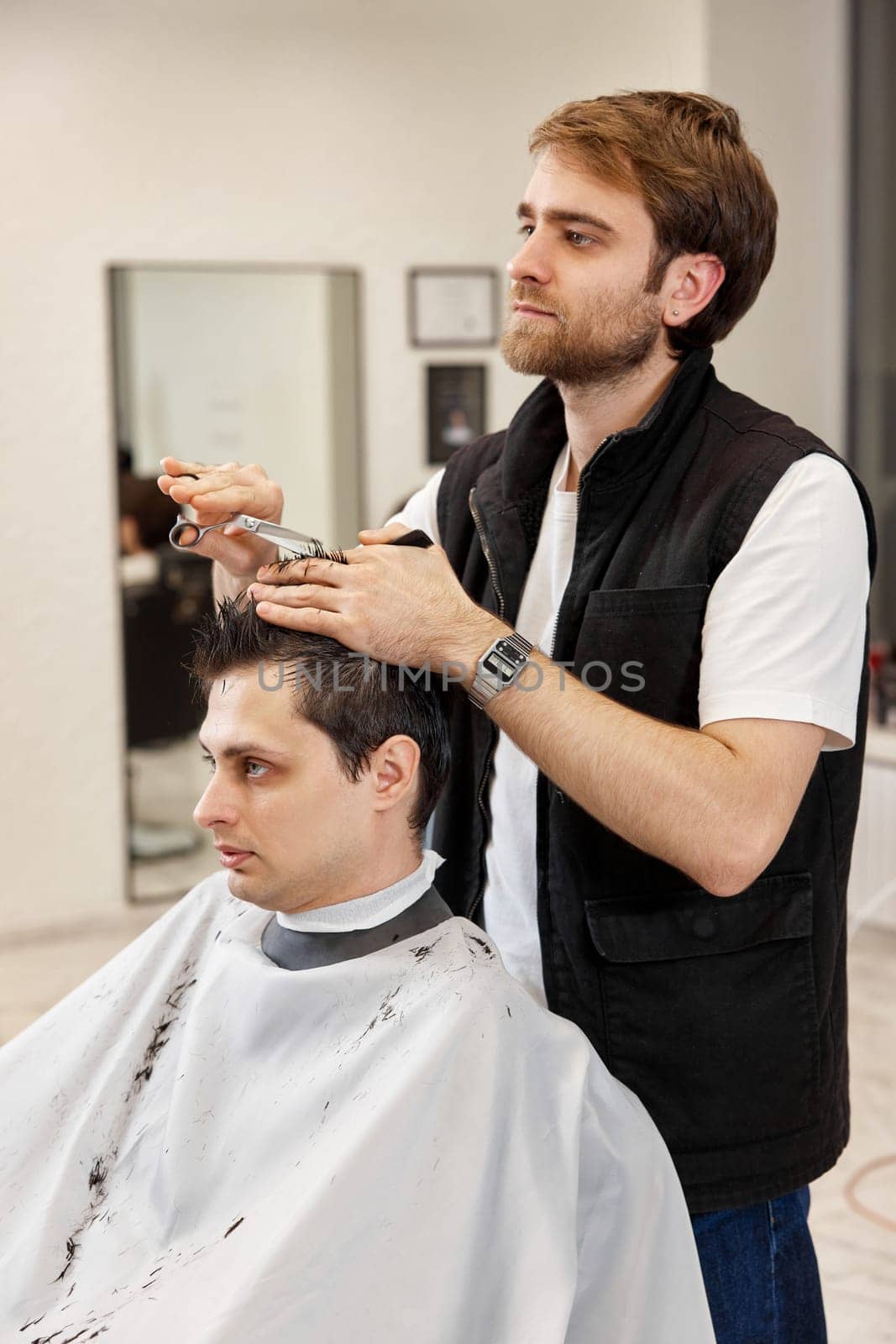 Professional bearded hairdresser does haircut for caucasian client man at barber shop.