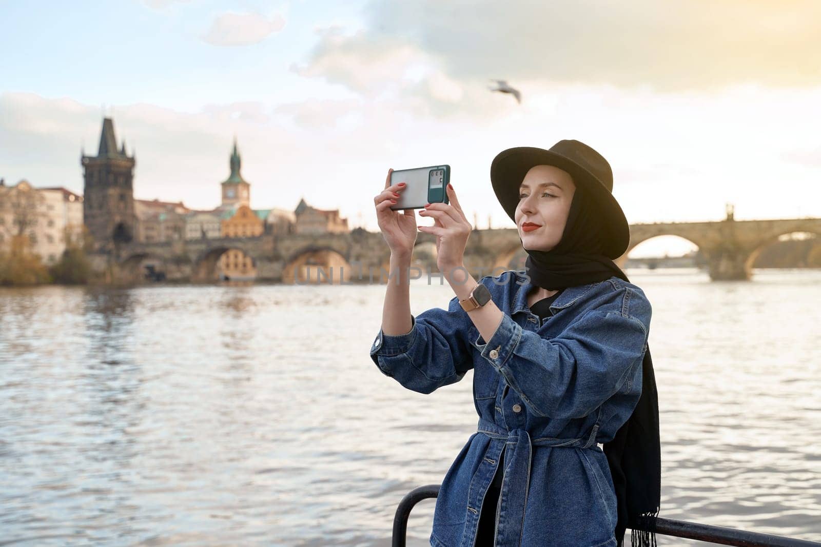 Stylish young woman traveller wearing black hat sitting taking pictures with her smartphone on Vltava river shore in Prague with Charles Bridge on background. by berezko
