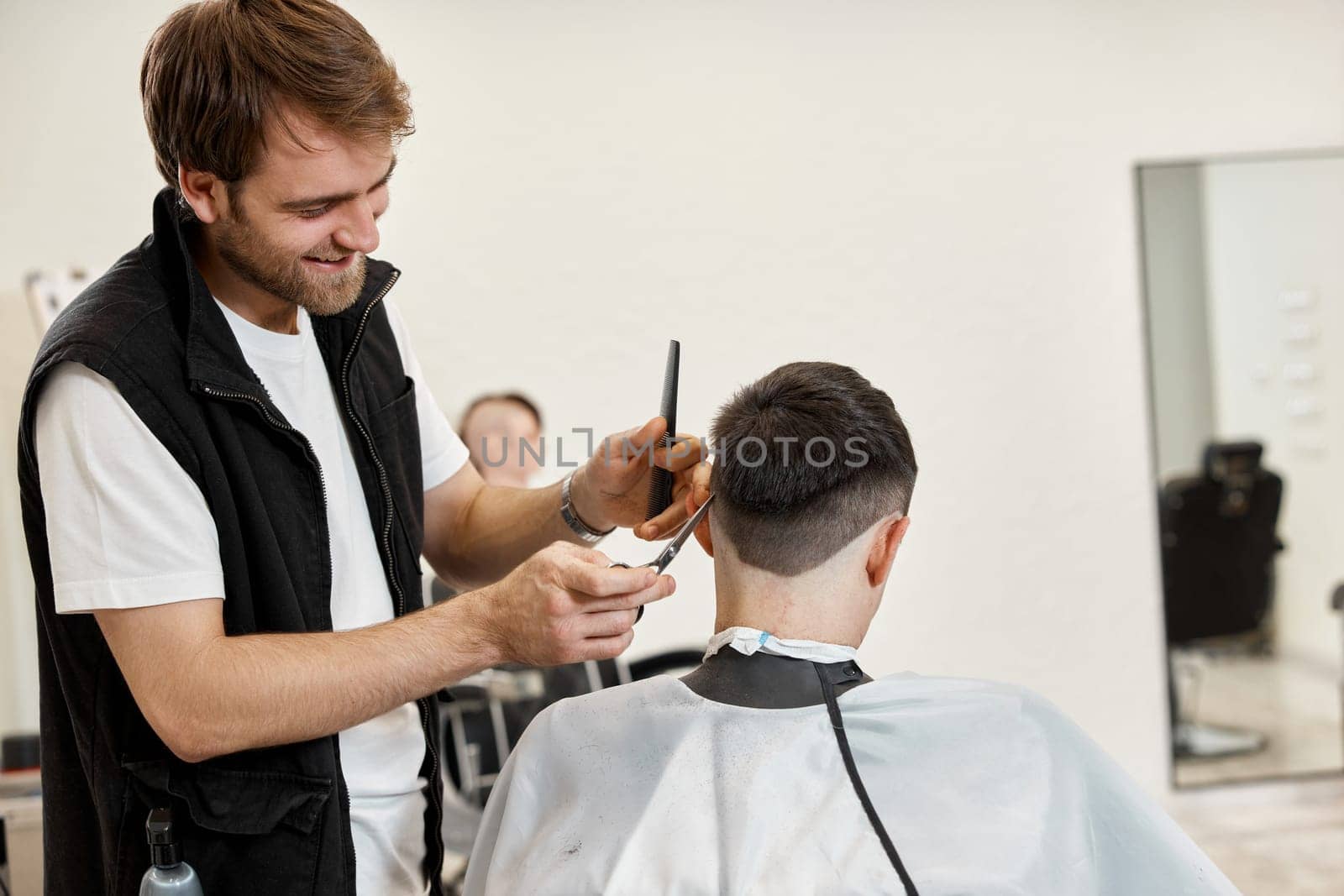 Professional hairdresser does haircut for caucasian bearded man using comb and scissors at barber shop.