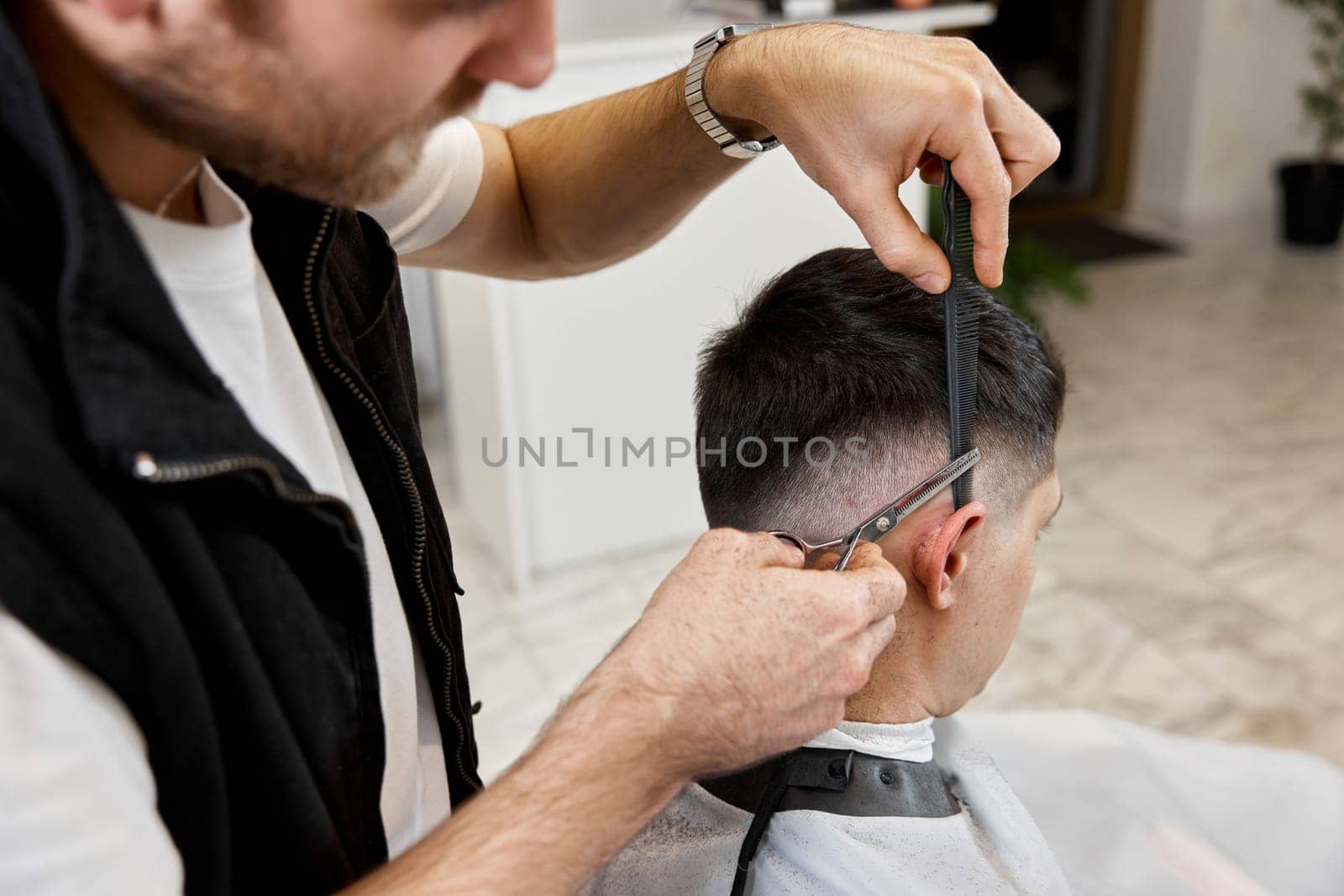 Professional hairdresser does haircut for caucasian bearded man using comb and scissors at barber shop.