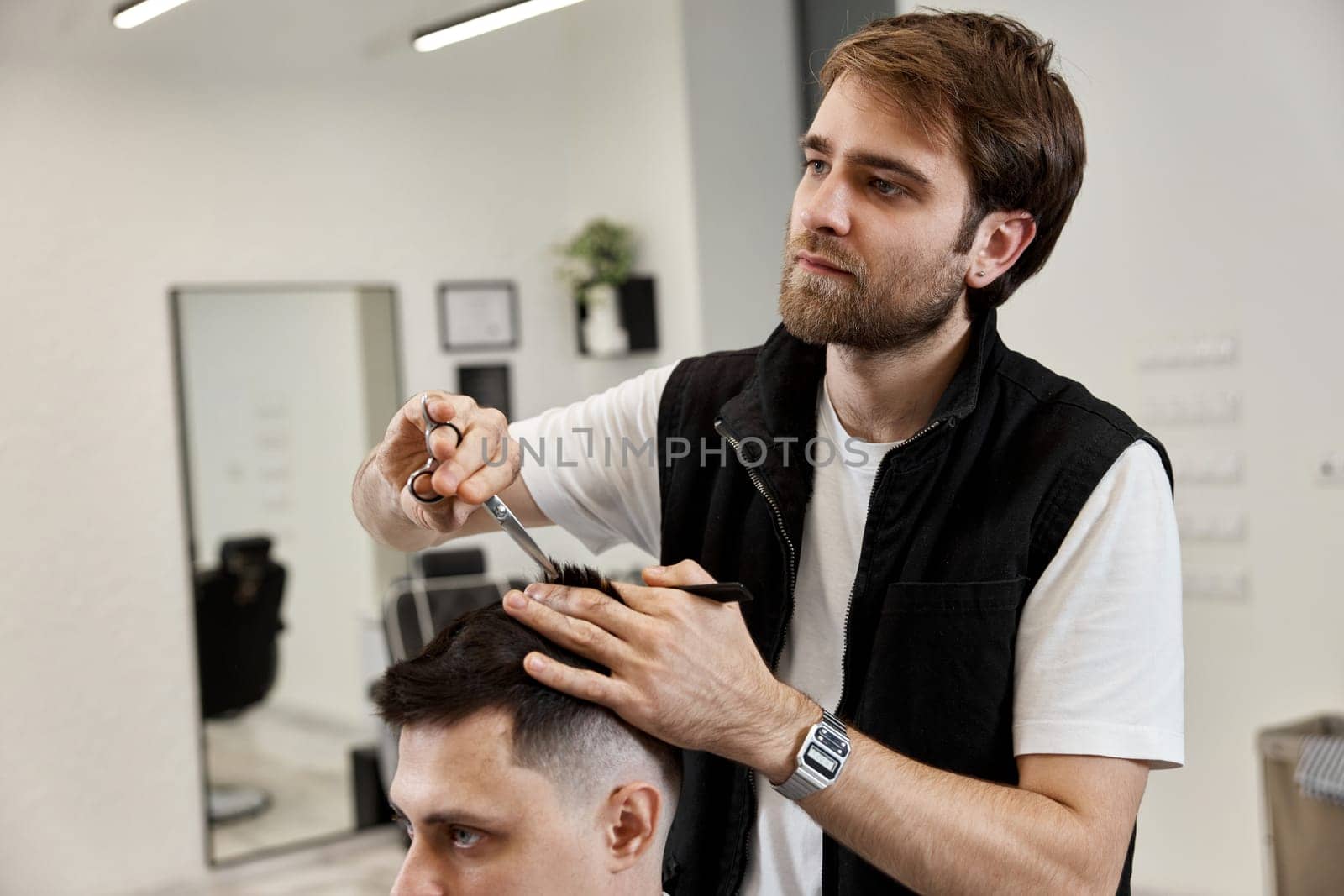 Professional hairdresser does haircut for caucasian bearded man using comb and scissors at barber shop.