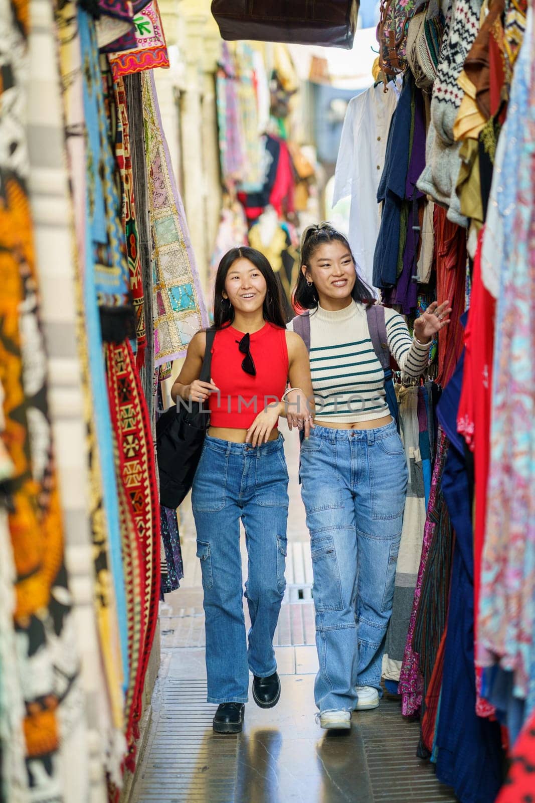 Full body of smiling young female friends in casual clothes walking along colorful clothing store on street market in city during summer vacation
