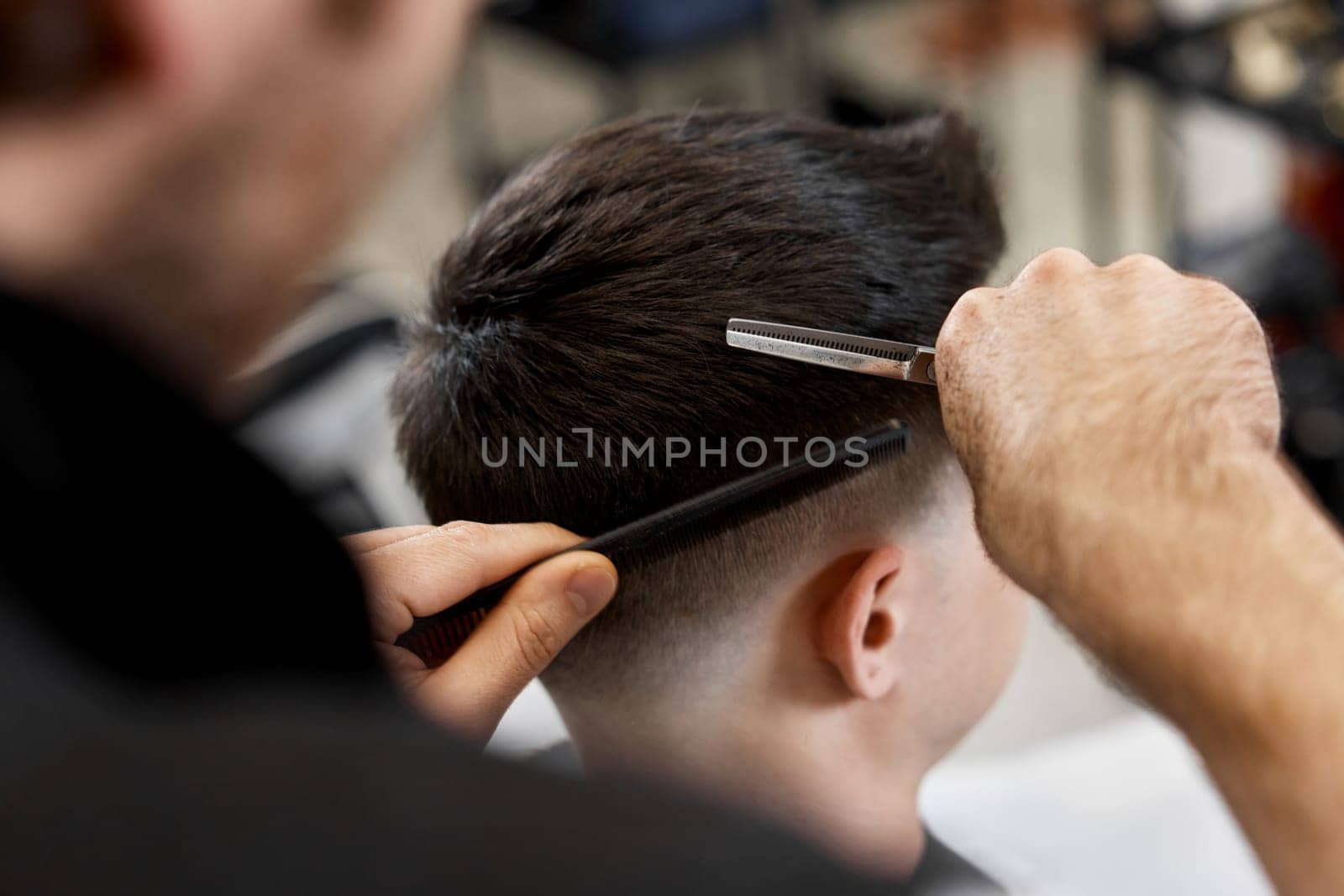 Professional hairdresser does haircut for caucasian bearded man using comb and scissors at barber shop.