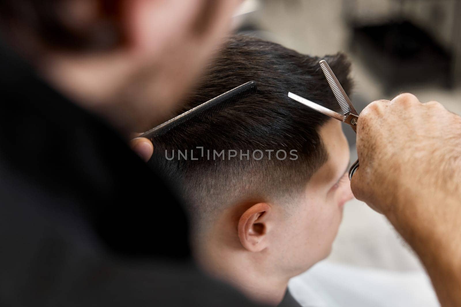 Professional hairdresser does haircut for caucasian bearded man using comb and scissors at barber shop.