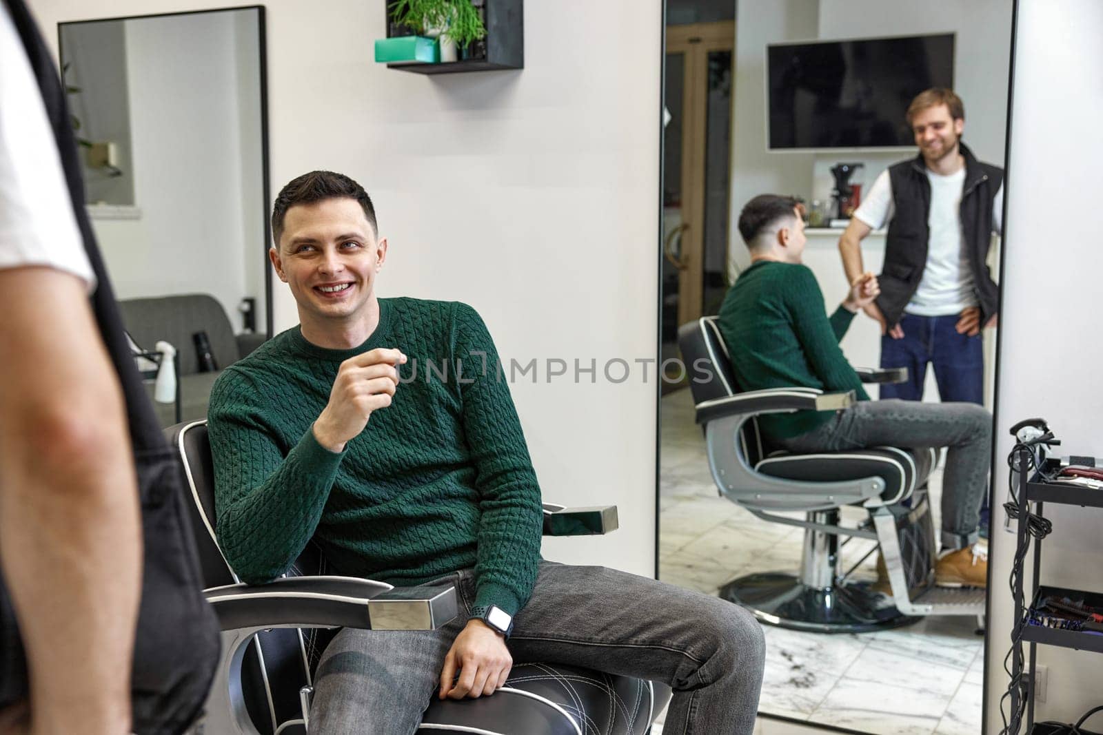 satisfied caucasian client man talking with barber while sitting in chair after haircut at barbershop
