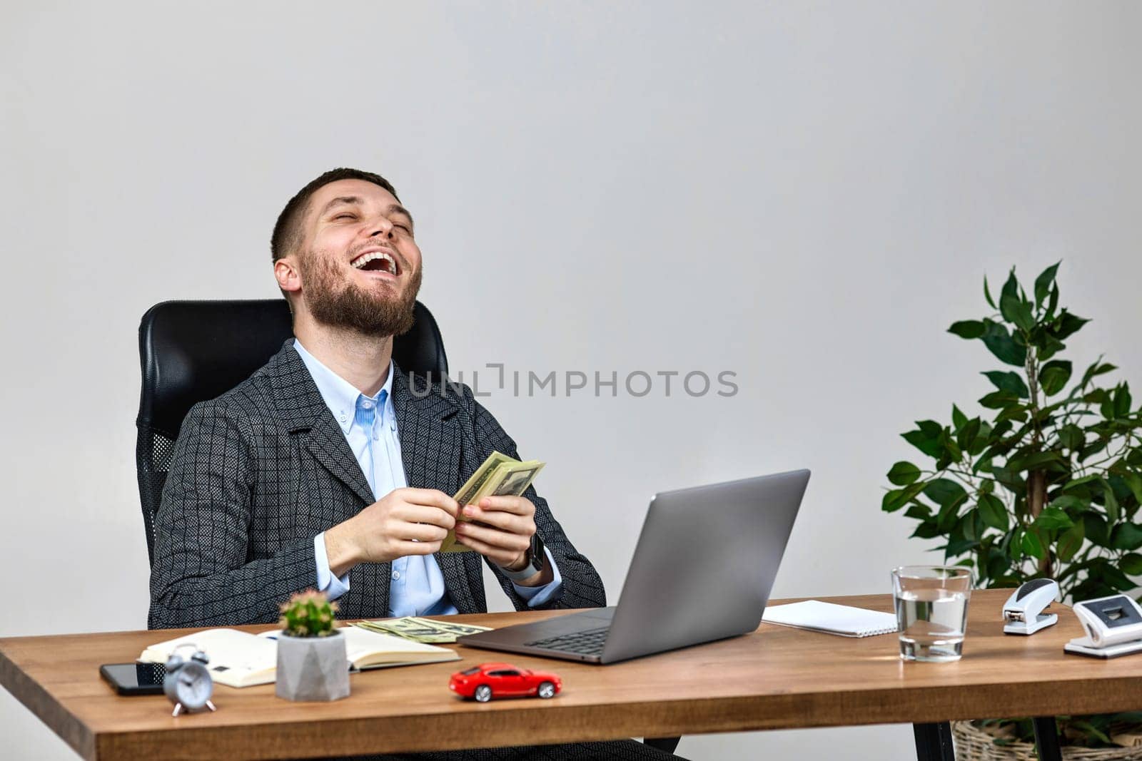 young bearded businessman working on laptop and counting dollars while sitting on chair at desk.