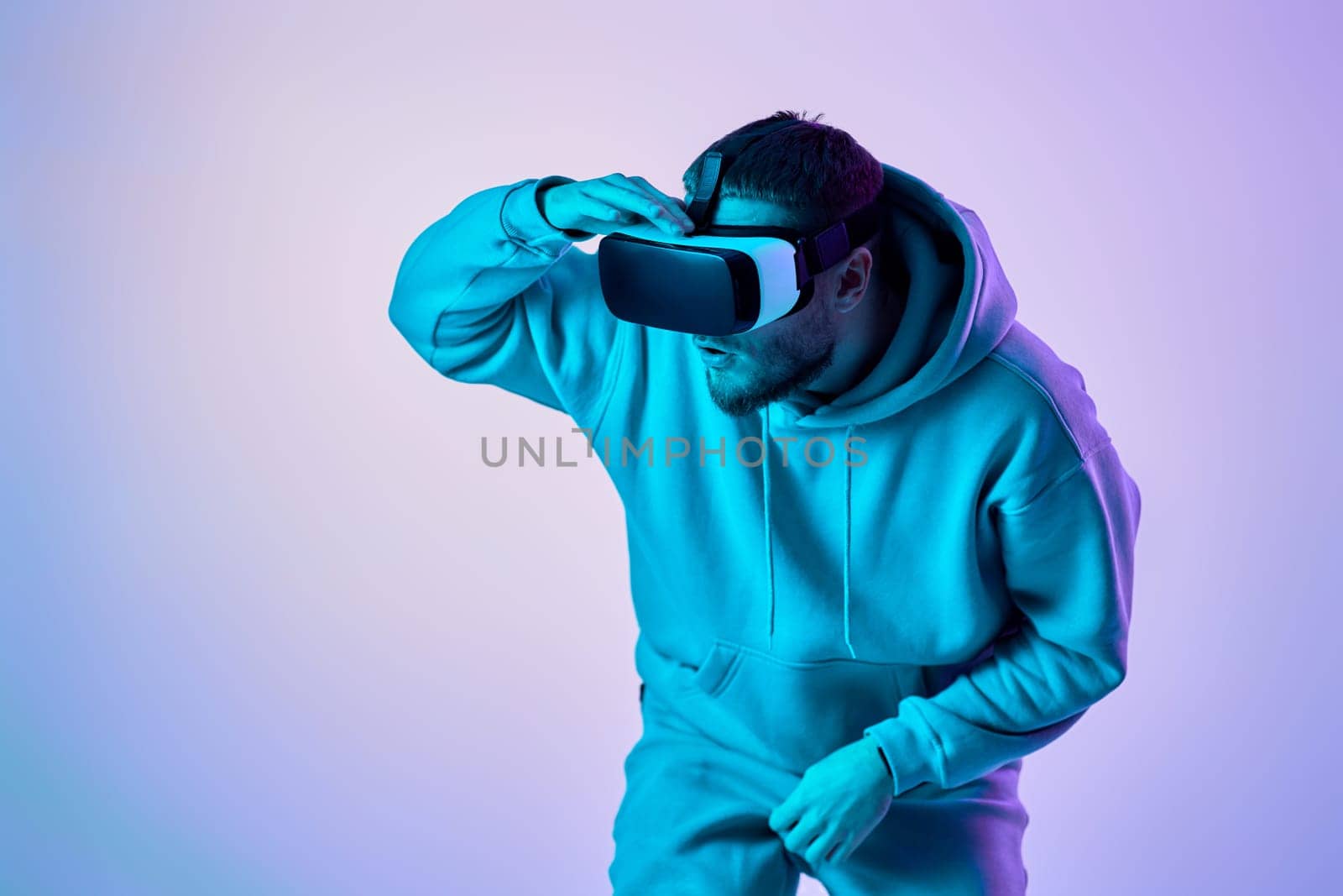 man in sweatshirt using virtual reality googles and playing video games on blue and pink neon background.