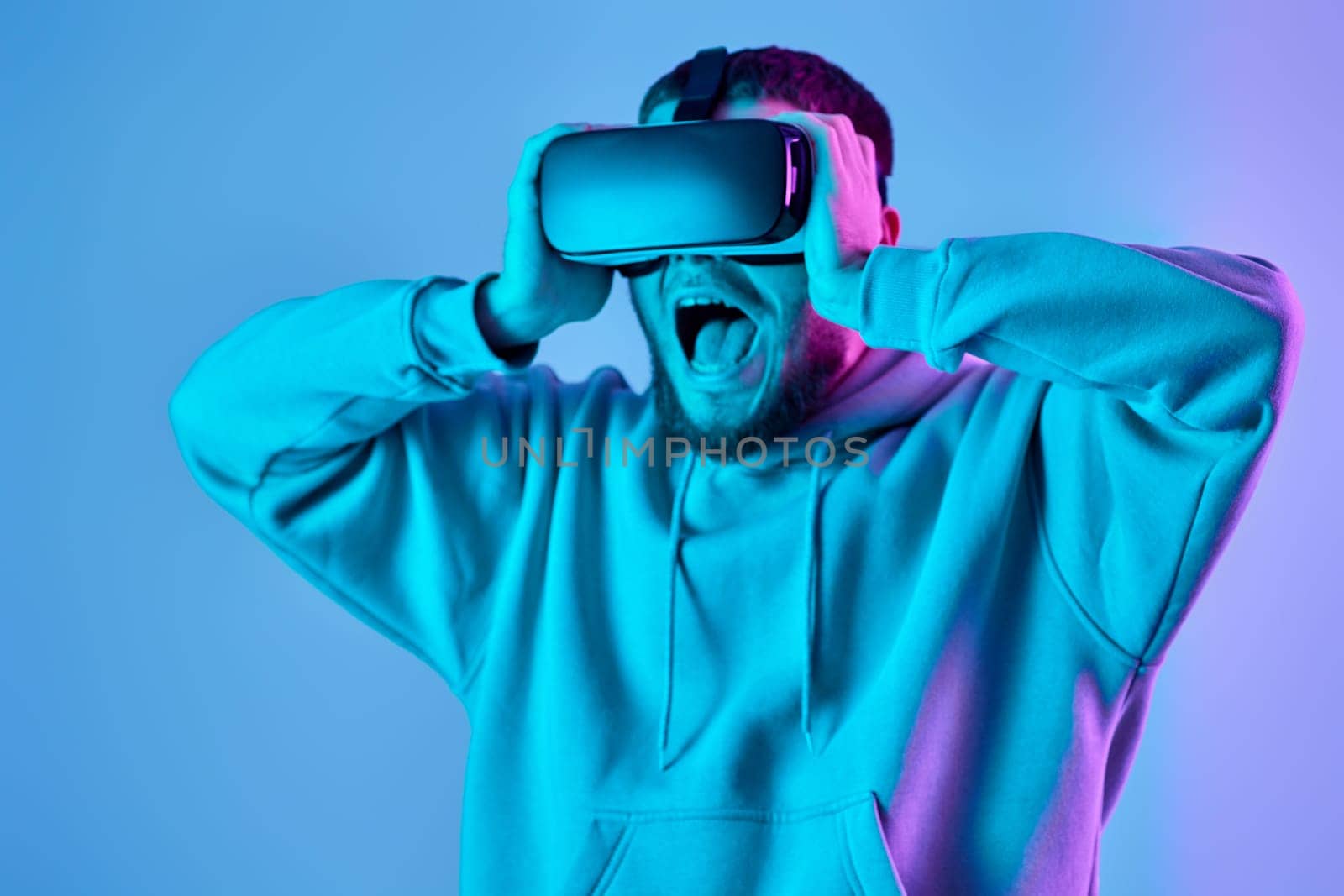 surprised man in sweatshirt using virtual reality glasses and playing video games on blue background. Neon lighting