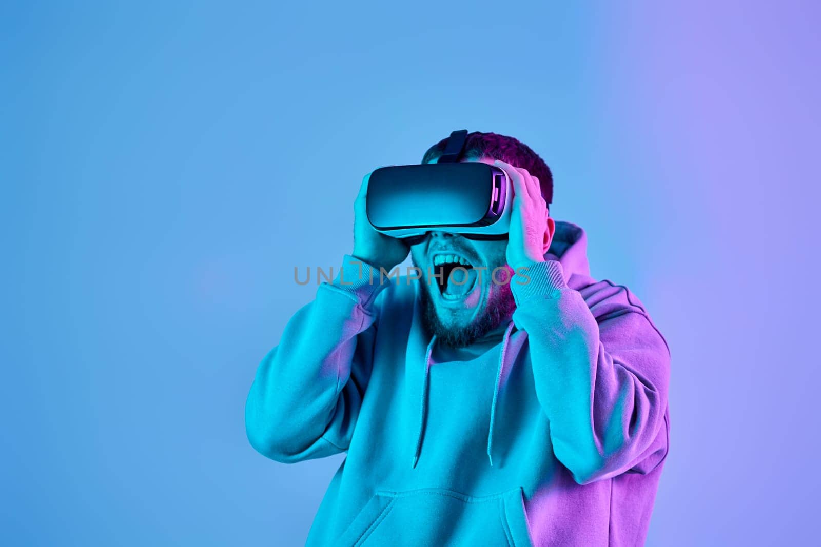 surprised man in sweatshirt using virtual reality glasses and playing video games on blue background. Neon lighting