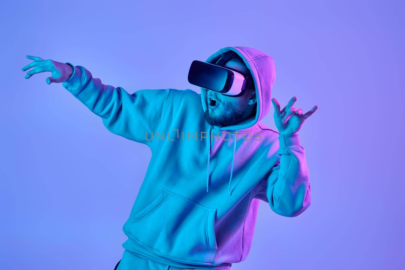 Excited man in sweatshirt using virtual reality glasses and playing video games on blue and pink neon background.