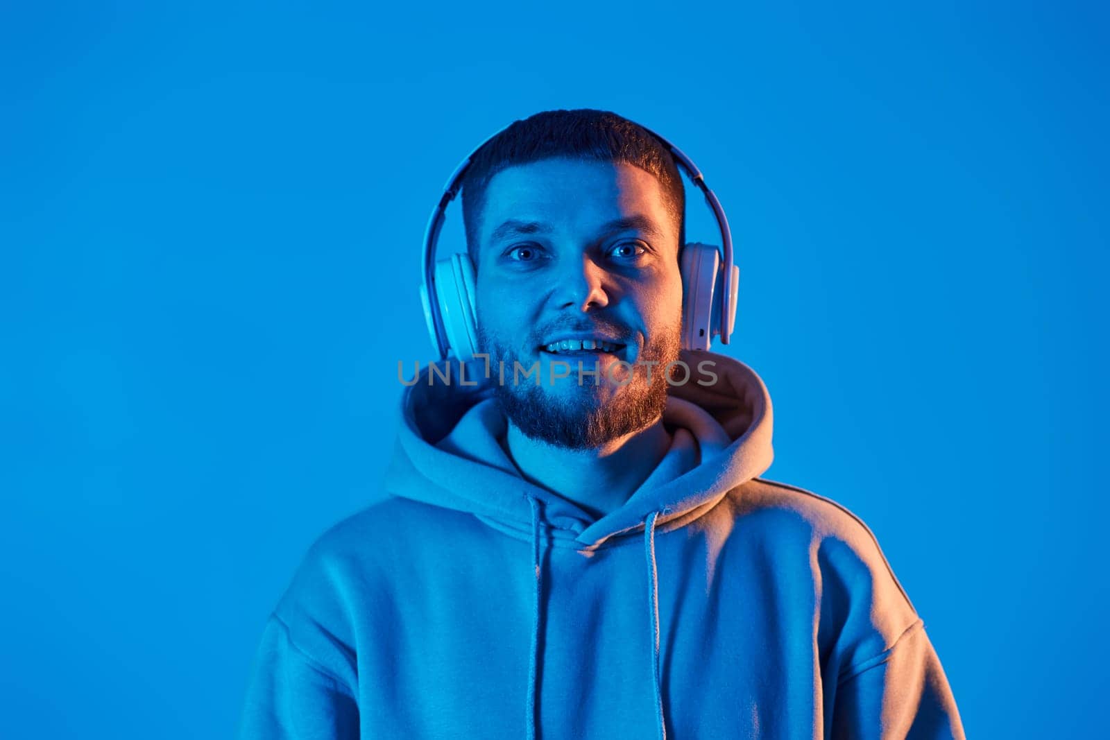 handsome bearded man with headphones in sweatshirt enjoying favorite music on blue neon background. Neon lighting