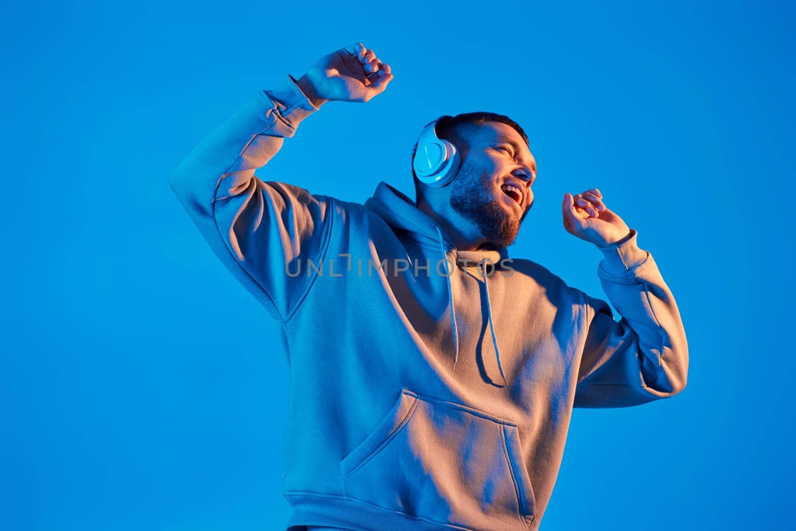 handsome bearded man with headphones in sweatshirt enjoying favorite music on blue neon background. Neon lighting