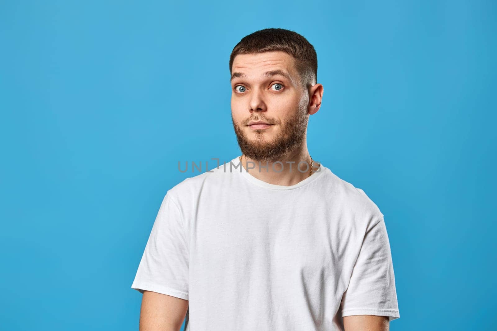 puzzled young bearded man in casual white t-shirt looking at the camera on blue gray background.