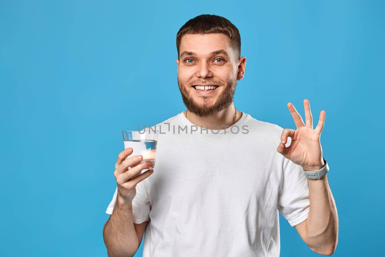 handsome bearded man in white t-shirt with drinking glass of water showing Ok gesture on blue background