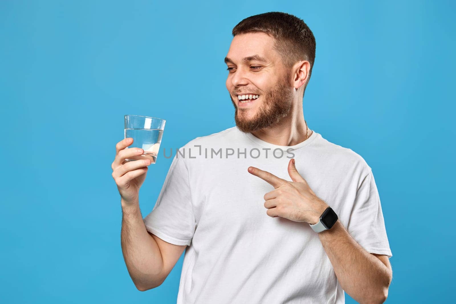 positive handsome man in white t-shirt showing drinking glass of water on blue background