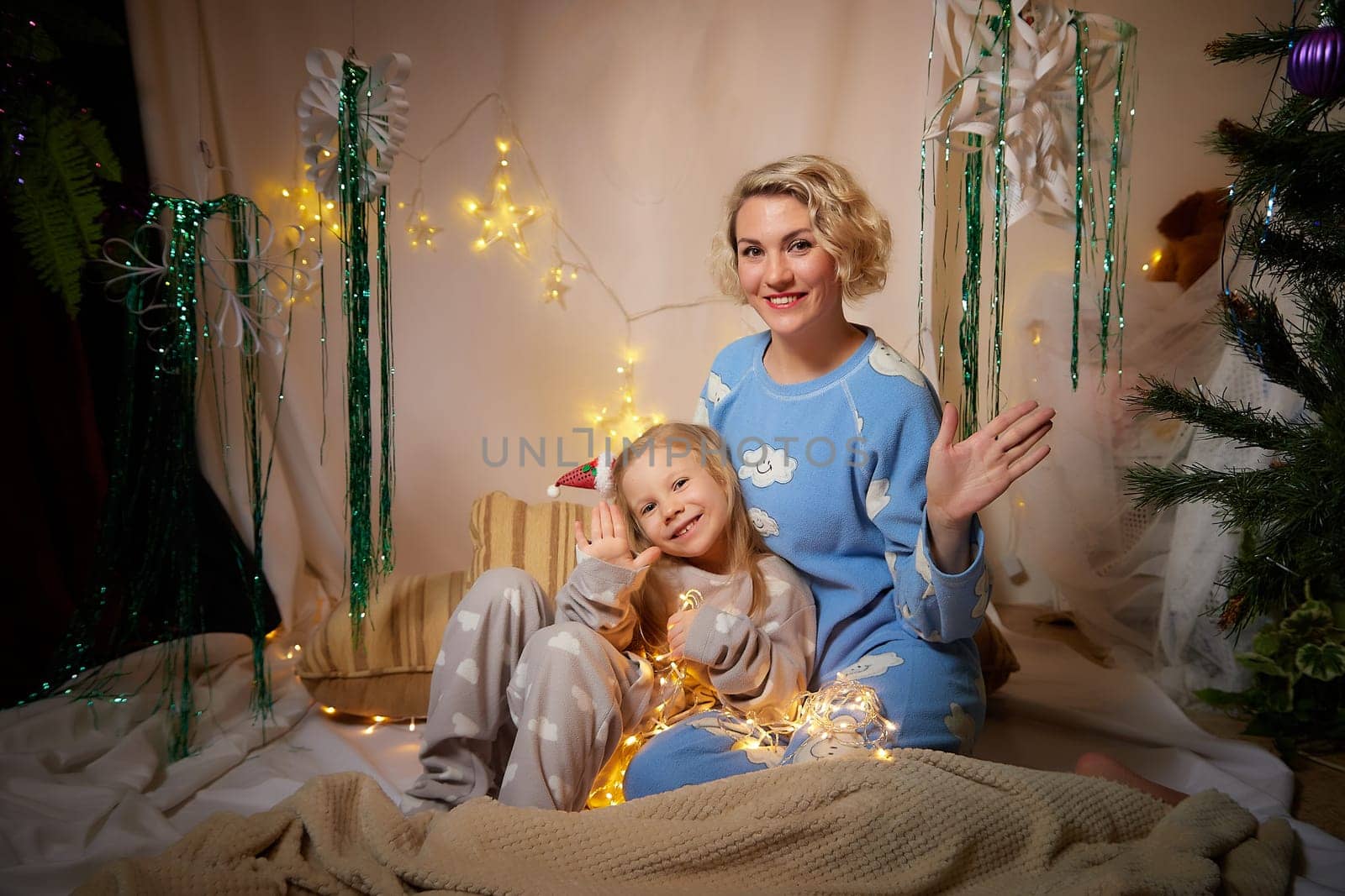Cute mother and daughter in pajamas having fun in the room with Christmas garlands and white background. The tradition of decorating the house for the holidays. Happy childhood and motherhood by keleny