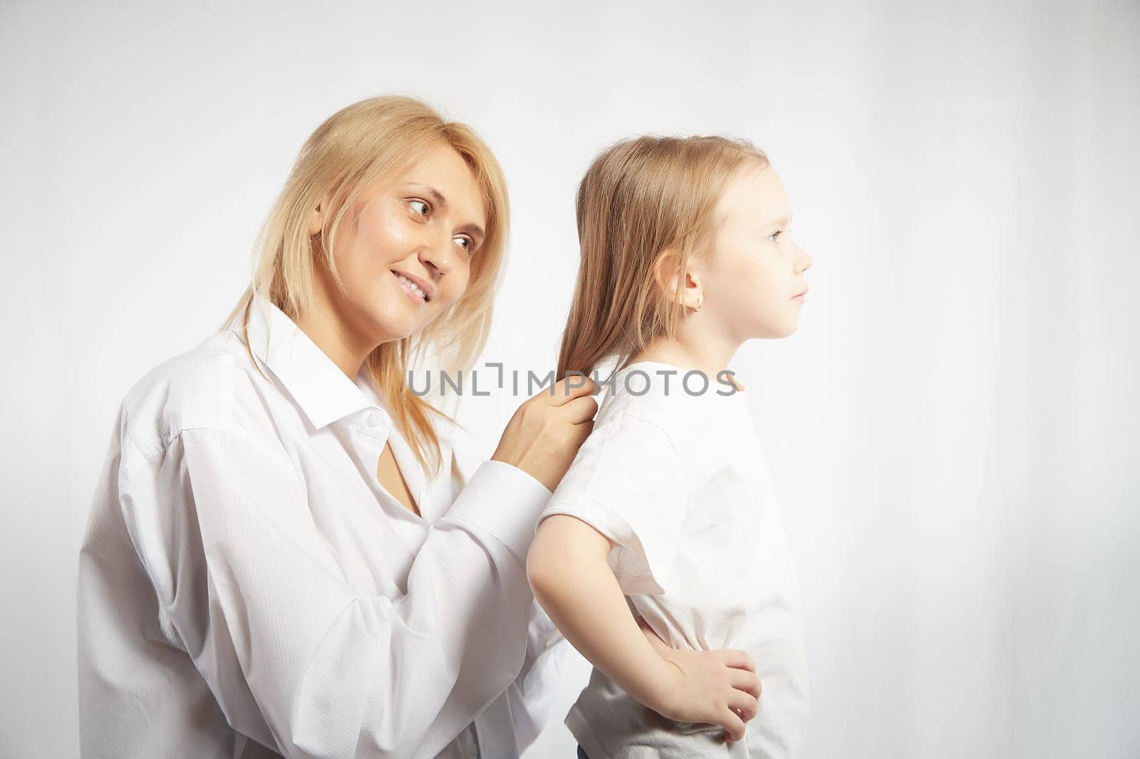 Close portrait of blonde mother and daughter where mom braids hair and makes ponytail and hairstyle for her girl on white background in studio. The concept of love, friendship, caring in the family by keleny