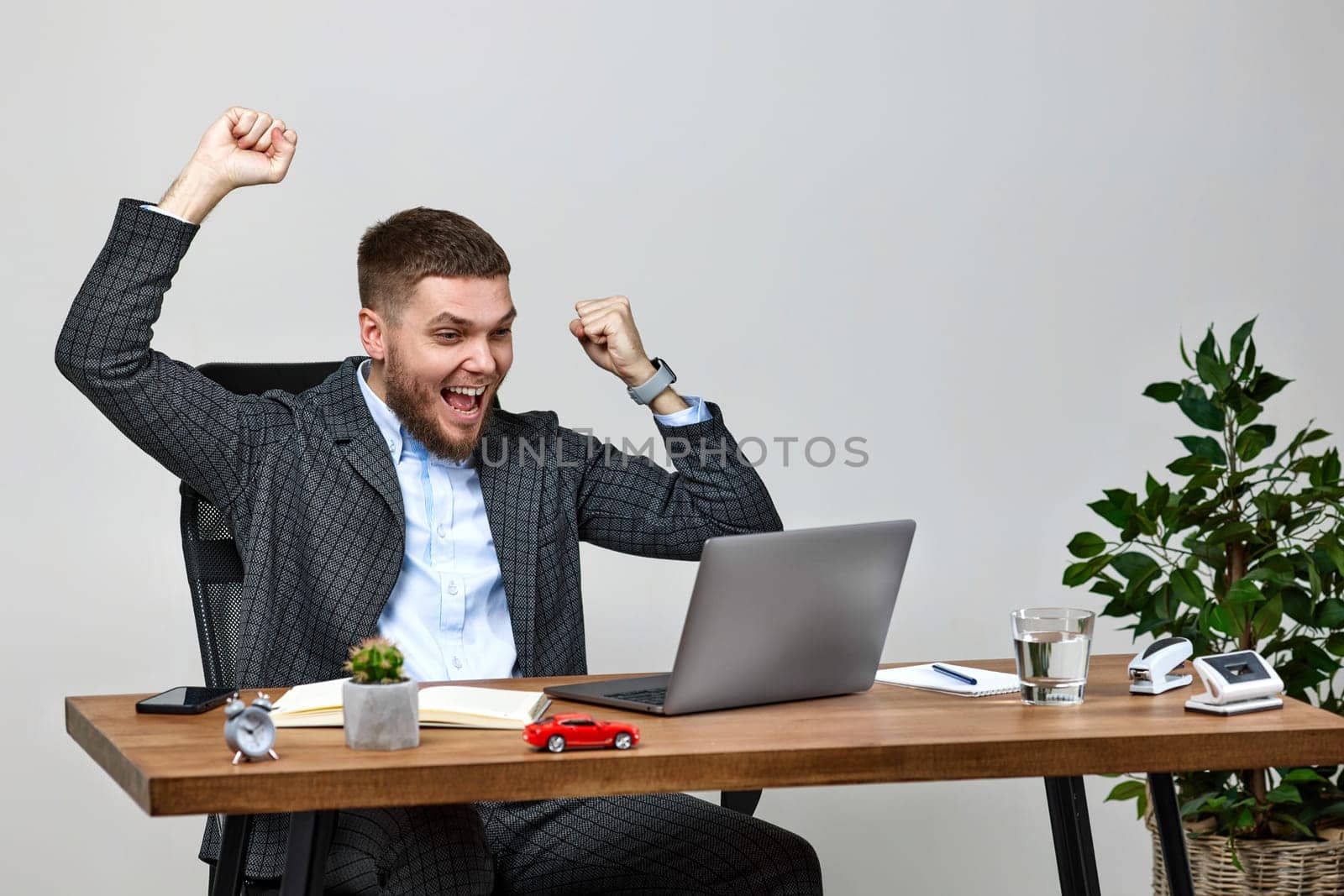 Satisfied young businessman gesturing emotionally and celebrating victory while sitting on chair at desk, using laptop pc computer