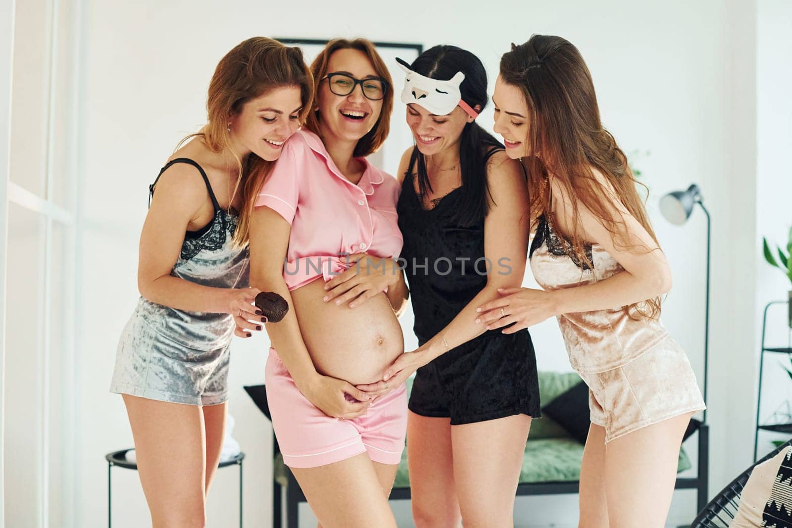 Cheerful young women with their pregnant friend in pajamas standing and have a party indoors at daytime together by Standret