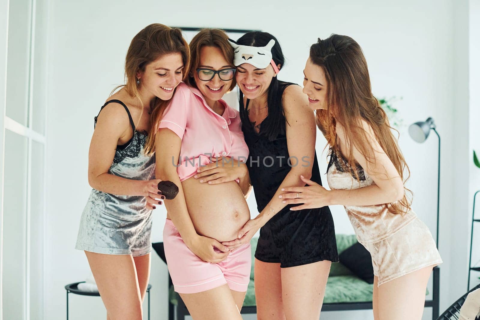 Cheerful young women with their pregnant friend in pajamas standing and have a party indoors at daytime together.