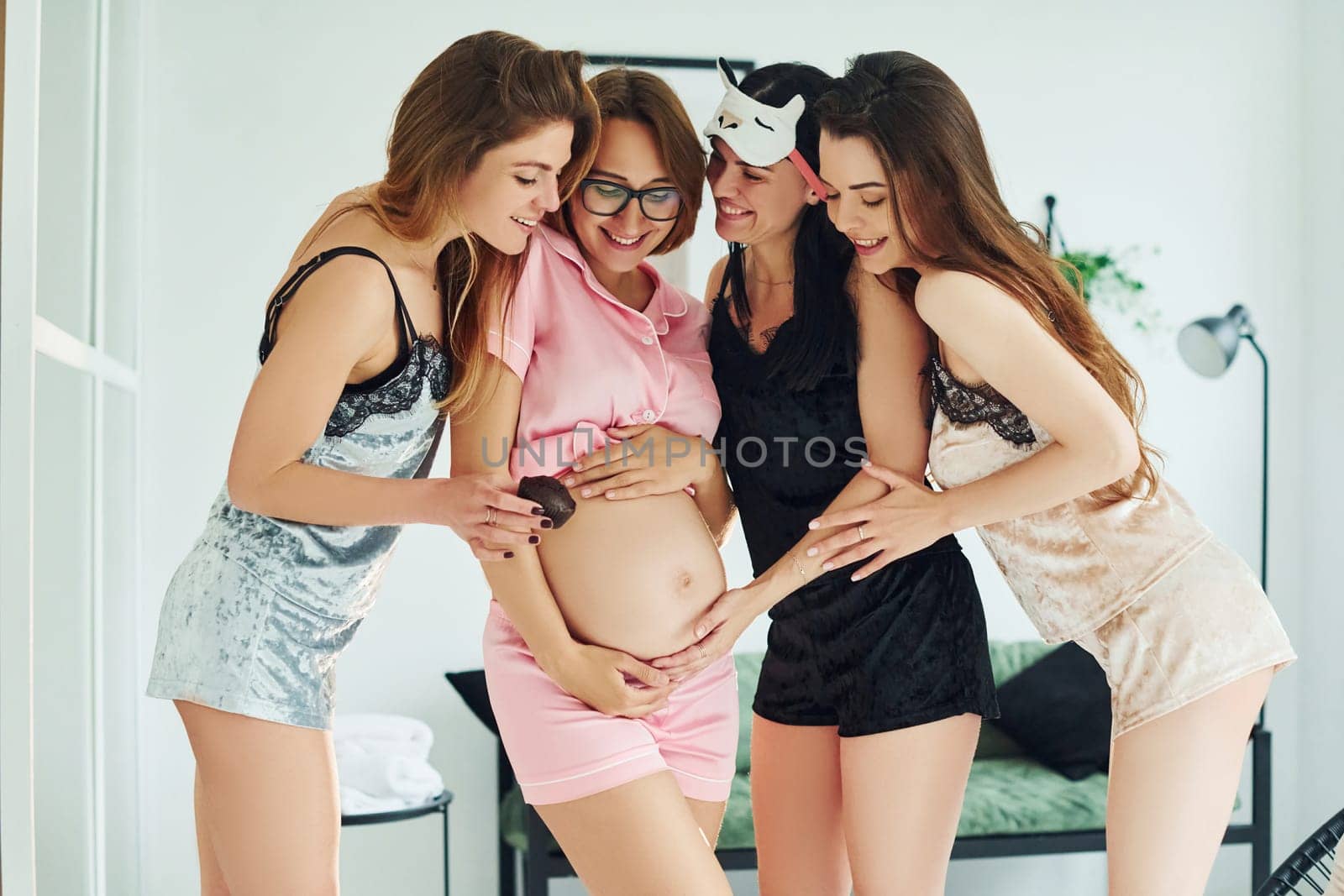 Cheerful young women with their pregnant friend in pajamas standing and have a party indoors at daytime together by Standret
