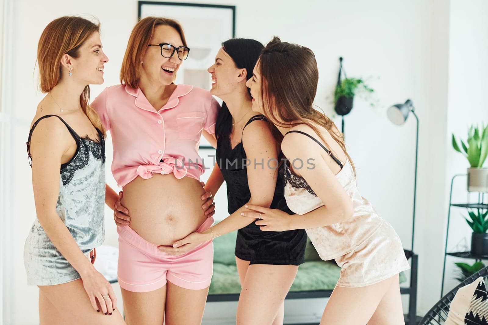 Cheerful young women with their pregnant friend in pajamas standing and have a party indoors at daytime together.
