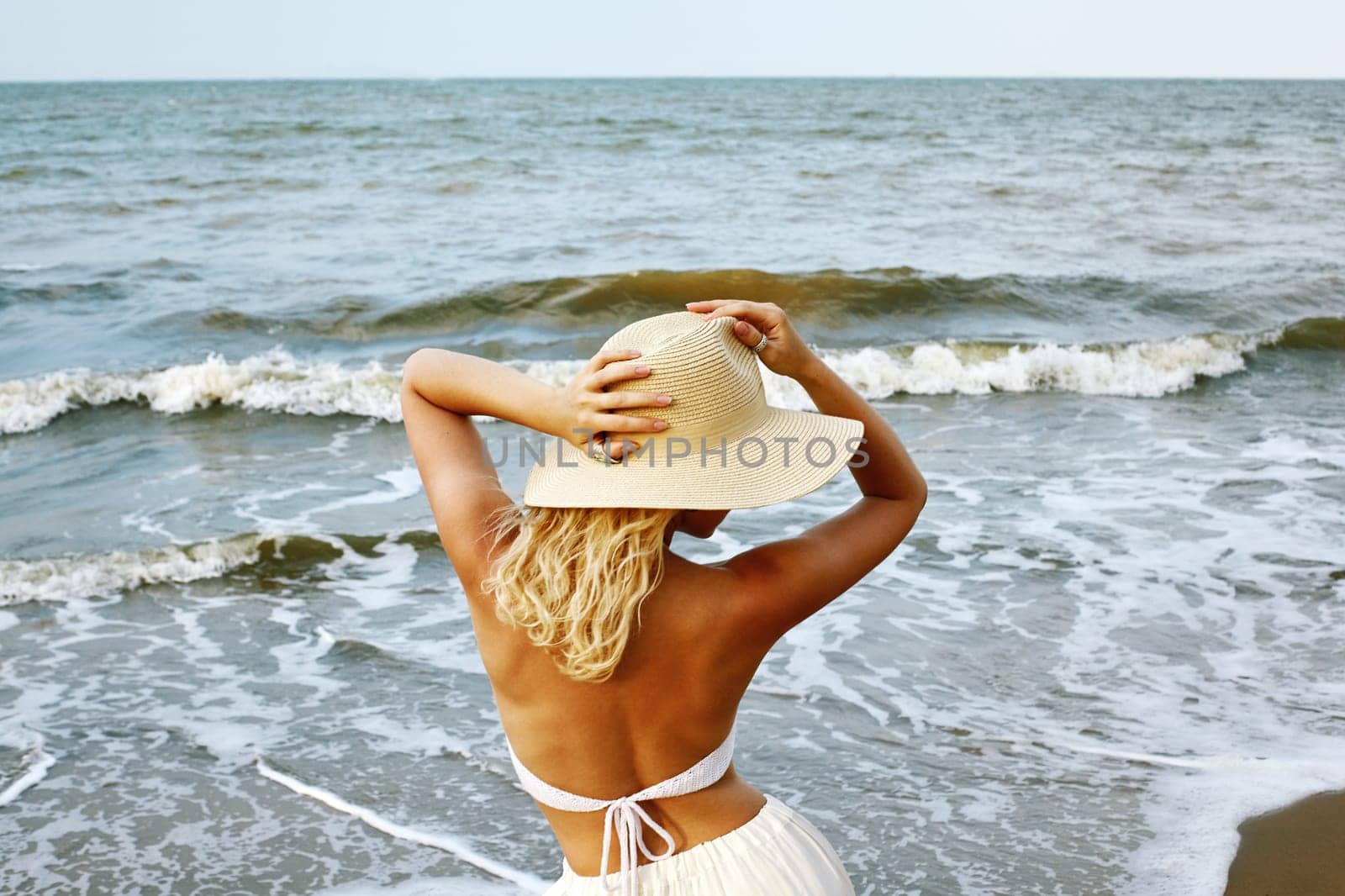 Blonde Woman in White Summer Style Standing at Sea and Holding Hat by MarinaFrost