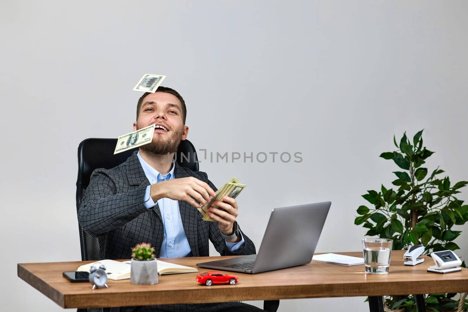 young bearded businessman working on laptop and throwing money while sitting on chair at desk. banknotes fly in the air