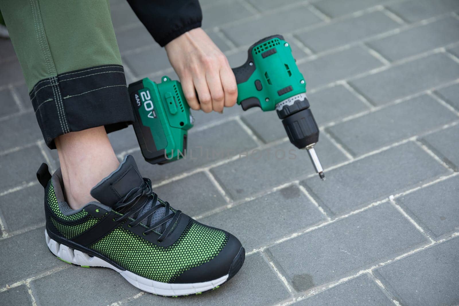 The girl is holding a cordless screwdriver and is dressed in boots and work pants. The background is made of laid rectangular paving stones.