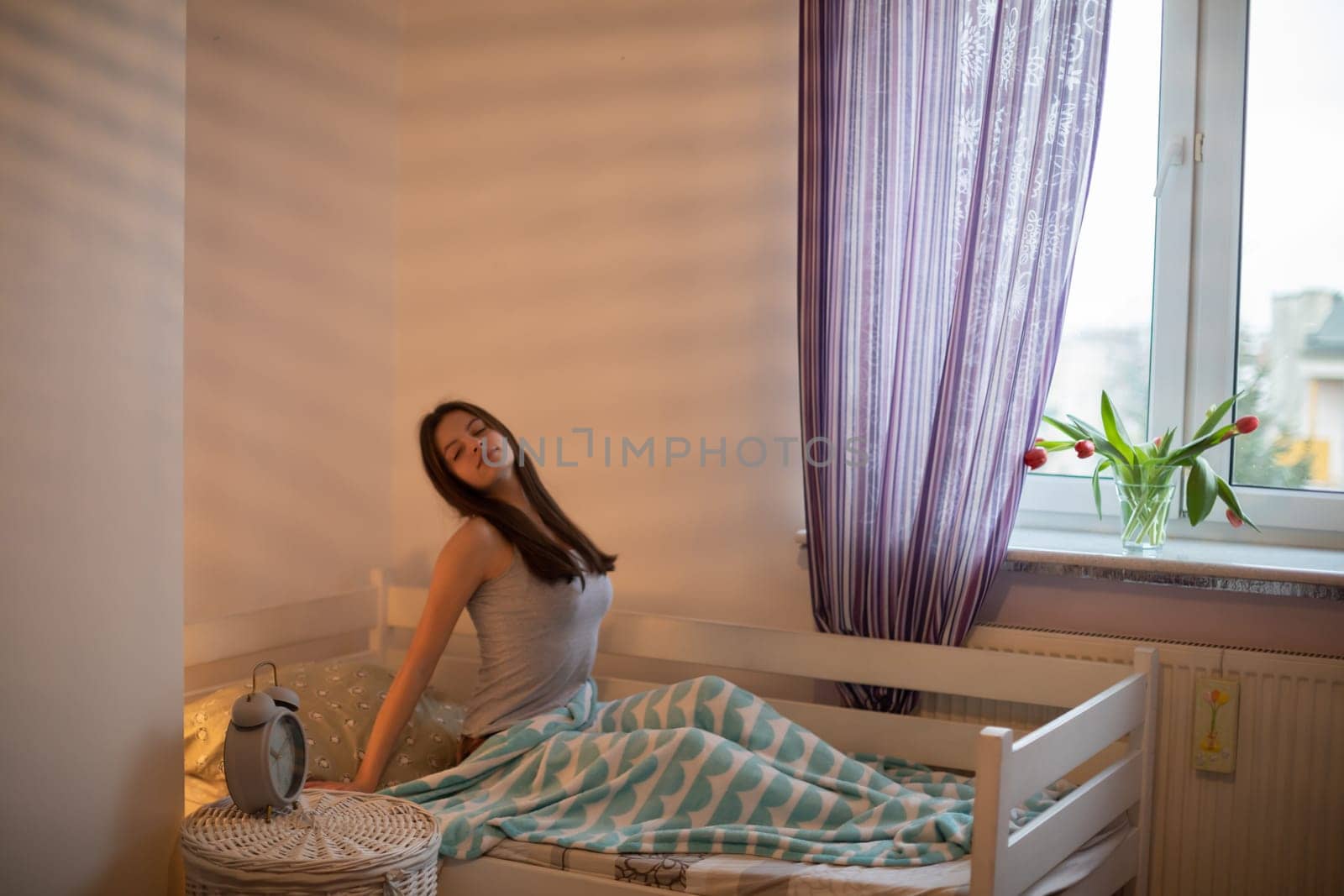 A girl with brown hair sits on her bed by the window and stretches after sleeping. A girl under a soft blanket.