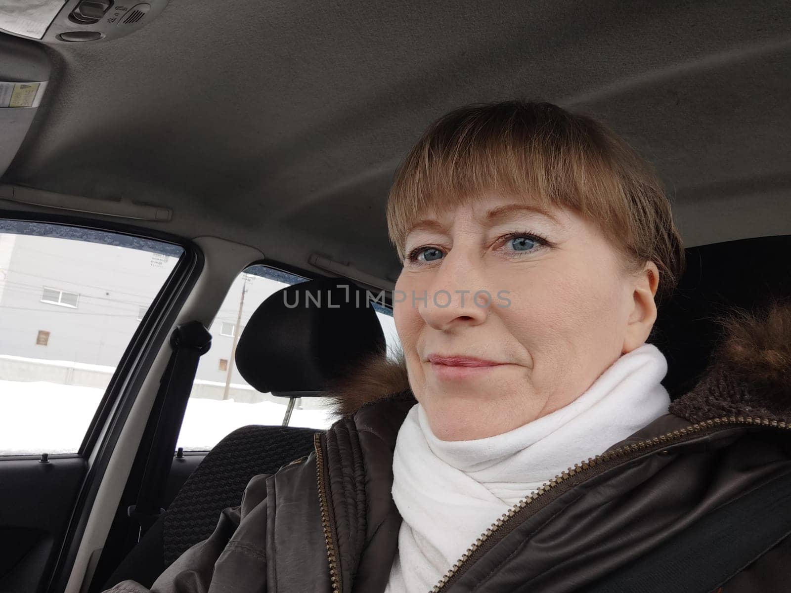 Portrait of adult cute funny blonde woman in warm clothes taking a selfie inside the car and driving it. The concept of pleasant trip in spring, autumn, winter